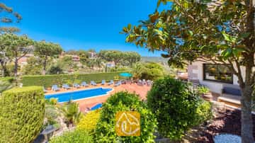 Außenterrasse der Villa Jaruco mit einem blauen Swimmingpool, gesäumt von gestreiften Sonnenliegen und umgeben von grünen Bäumen unter einem klaren blauen Himmel