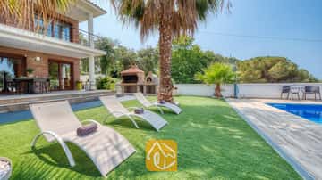 Sonniger Garten der Villa Marcella mit weißen Sonnenliegen auf Kunstrasen, einer Palme und einem blau gefliesten Swimmingpool neben der Ziegelstein-Außenfassade des Hauses.