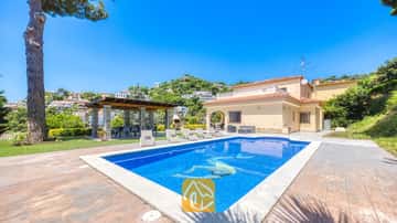 Der Außenpoolbereich der Villa Paris mit klarem blauen Wasser, Sonnenliegen auf der gefliesten Terrasse und einer überdachten Pergola direkt am zweistöckigen, beigen Haus.