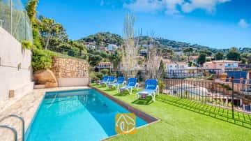 Privater Pool mit blauen Liegen auf einer grünen Kunstrasenterrasse in der Villa Donna, mit Blick auf Hanghäuser unter einem klaren blauen Himmel.