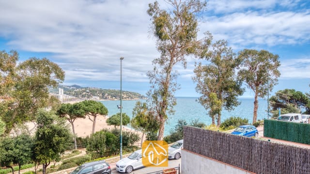 Blick von der Casa Costa auf hohe Bäume, einen Sandstrand und das blaue Meer unter einem leicht bewölkten Himmel.