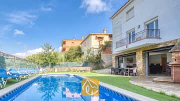 Privater Pool und Terrasse der Villa Nicky mit blauen Liegestühlen auf Kunstrasen neben einer zweistöckigen weißen Villa mit Steinsäulen und Glas Schiebetüren.