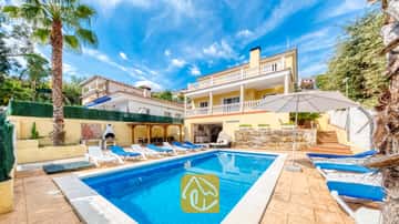 Der private Swimmingpool der Villa Ashley ist umgeben von blauen Sonnenliegen, einem weißen Sonnenschirm und einem zweistöckigen gelben Gebäude mit weißen Balkongeländern.