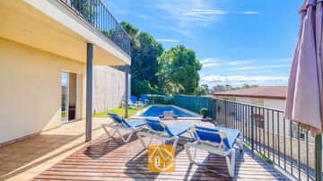 Sonnige Holzterrasse der Villa Mauri mit blauen Liegestühlen, einem darunter liegenden Swimmingpool und einem überdachten Terrassenbereich vor einem blauen, wolkenlosen Himmel