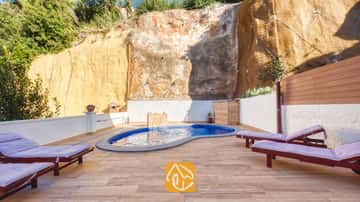 Der private Pool der Villa Blanca mit blauen Mosaikfliesen liegt auf einer gefliesten Terrasse, die von einer hohen Felswand eingerahmt wird, und lädt mit Holzliegen und violetten Kissen zum Entspannen ein.