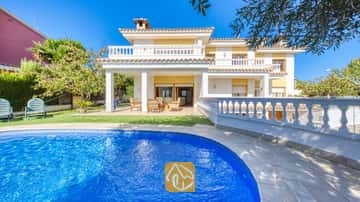 Ein geschwungener, blau gefliester Swimmingpool im Vordergrund, dahinter die Villa Baileys mit ihrer weißen Außenfassade, gelben Wänden und gefliesten Balkonen unter einem klaren Himmel.