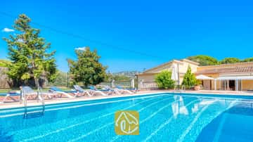 Außenpool der Villa Miro mit blau-weißen Sonnenliegen auf einer Terrasse aus Terrakotta. Grüne Bäume und ein gelbes Gebäude unter einem strahlend blauen Himmel.