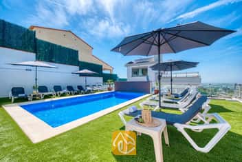 Privater Pool in der Villa Sandy Beach, umgeben von Kunstrasen, grauen Sonnenliegen mit weißem Rahmen und großen Sonnenschirmen unter einem strahlend blauen Himmel.
