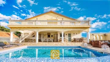 Außenansicht der Villa Madonna mit einem Swimmingpool, der ein Delfin-Mosaikmuster aufweist, einer schattigen Veranda, einer steinverkleideten Terrasse und gelben Außenwänden unter einem blauen Himmel.