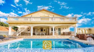 Außenansicht der Villa Madonna mit einem Swimmingpool, der ein Delfin-Mosaikmuster aufweist, einer schattigen Veranda, einer steinverkleideten Terrasse und gelben Außenwänden unter einem blauen Himmel.