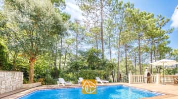 Der private Pool der Villa Esmee lädt mit weißen Sonnenliegen auf der gefliesten Terrasse, umgeben von hohen Pinien und einem Steingrill, zum Entspannen ein.