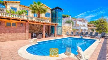Privater Pool und Terrasse in der Villa Dolce Vita mit gläsernem Aufzugsschacht, Backsteinwänden und mehreren Sonnenliegen unter blauen Himmel.