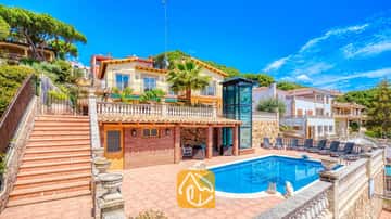 Außenansicht der Villa Dolce Vita mit Swimmingpool, einer Terrasse aus rotem Backstein, einer Außentreppe und einem modernen Aufzugsschacht aus Glas vor einem blauen Himmel.