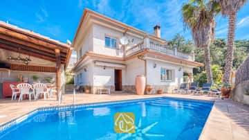 Außenansicht der Villa Valeria mit einem gefliesten Mosaik-Swimmingpool, einer zweistöckigen, weiß gestrichenen Villa mit Steinelementen, einer überdachten Terrasse mit weißen Stühlen und hohen Palmen.