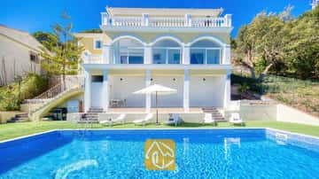 Außenbereich der Villa Sunrise mit einem rechteckigen, blau gefliesten Swimmingpool, weißen Sonnenliegen und einer gelben, mehrstöckigen Villa mit Bogen-Glastüren unter klarem Himmel.