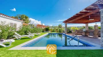 Außenpool und überdachte Terrasse der Villa Palmera mit weißen Plastiksonnenliegen auf Kunstrasen und einer Holzpergola mit Korbmöbeln.
