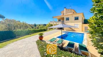 Außenansicht der Villa Holiday mit dem gelben zweistöckigen Haus, dem Swimmingpool mit Delphin-Mosaik und grünen Sonnenliegen unter einem grauen Sonnenschirm.