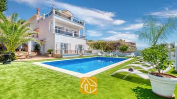 Privater Pool und weiße Sonnenliegen auf einer grünen Rasenfläche an der Villa Maribel in Spanien, mit einer mehrstöckigen weißen Villa mit Balkonen und Palmen.