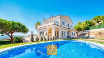 Privater Swimmingpool im Vordergrund mit der Villa Ibiza, einem zweistöckigen, cremefarbenen Haus, grünem Rasen und Kakteen in Töpfen unter einem strahlend blauen Himmel.