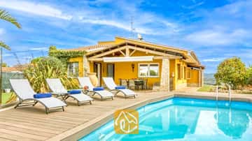 Privater Pool mit Holzterrasse und weißen Sonnenliegen an der Villa Soleia in Spanien, mit einer gelben Außenfassade der Villa und blauem Himmel.